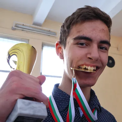 Момче, държащо златна купа, е захапал златен медал от състезание по физика.